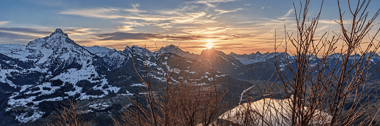 Verschneite Schweizer Alpen im Abendlicht mit leuchtendem Sonnenuntergang, markanten Berggipfeln und kahlen Ästen im Vordergrund.
