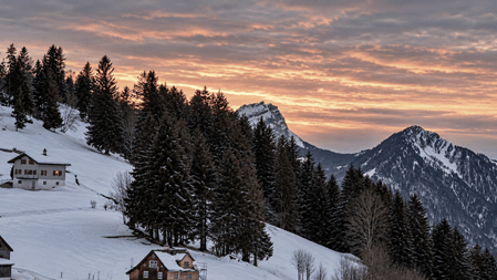 Verschneite Schweizer Berglandschaft bei Sonnenuntergang mit dunklen Tannen, verstreuten Berghäusern und markanten Gipfeln vor einem orangefarbenen, wolkenverhangenen Himmel.