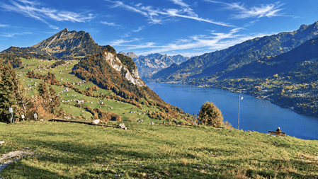 Aussichtspunkt mit grüner Wiese und Sitzbank über einem tiefblauen Bergsee, umgeben von herbstlich gefärbten Berghängen, verstreuten Häusern und einem eindrucksvollen Alpenpanorama unter blauem Himmel mit Wolken.