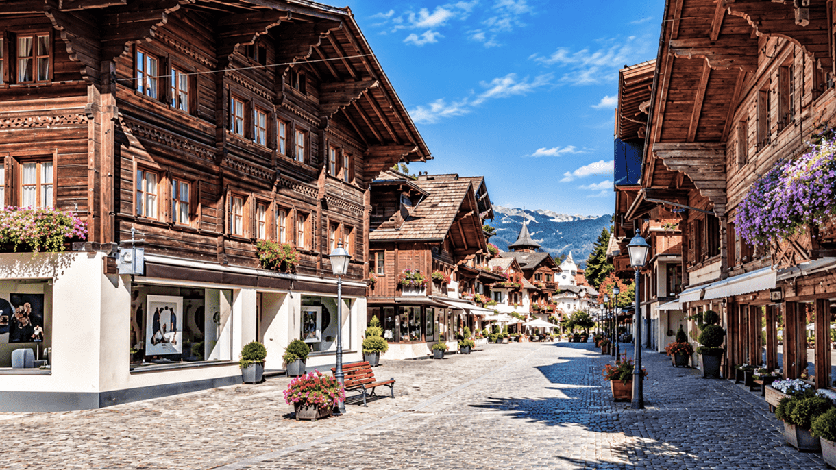 Gstaad: Historische Chalets mit Blumenschmuck säumen die gepflasterte Dorfstrasse, im Hintergrund die Schweizer Alpen unter blauem Himmel.