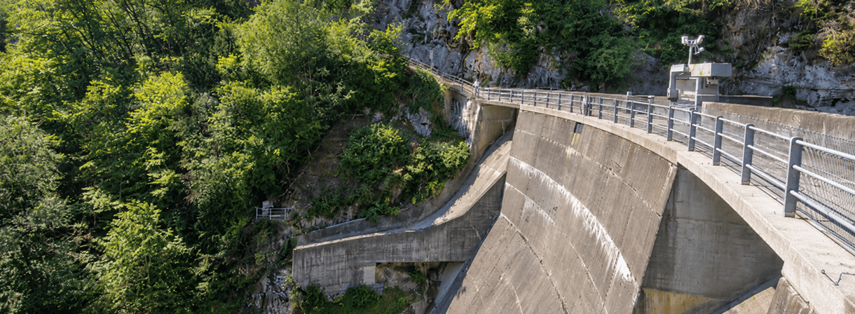 Gebogene Betonstaumauer mit begehbarem Geländer und technischem Kontrollgebäude in einer dicht bewaldeten Schlucht. Sonnenlicht hebt die Felswände, die grüne Vegetation und die Struktur der Staumauer hervor.