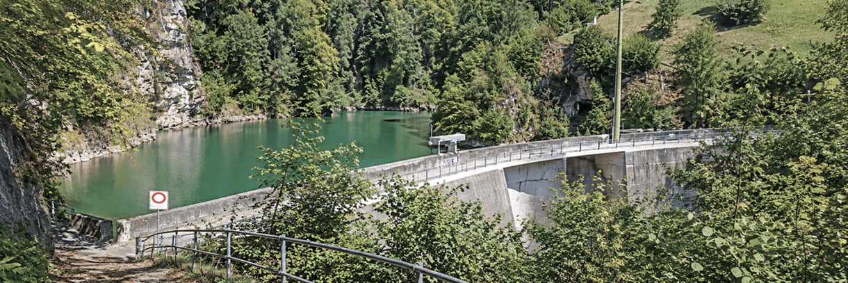Gebogene Betonstaumauer mit Geländer und türkisgrünem Stausee in einer bewaldeten Felsenschlucht, aufgenommen von einem Weg mit Metallgeländer im Vordergrund.