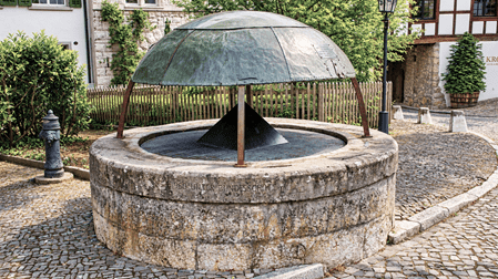 Historischer Sodbrunnen in Regensberg mit rundem Natursteinbecken und schützendem Metalldach. Der mittelalterliche Brunnen liegt auf einem gepflasterten Platz, umgeben von traditionellen Fachwerkhäusern, Steinmauern und gepflegten Grünanlagen.