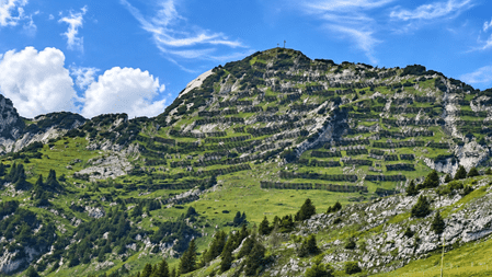 Terrassierter grüner Berghang mit steinigen Felsformationen, schmalen Wanderwegen und Gipfelkreuz unter blauem Himmel mit weissen Wolken.