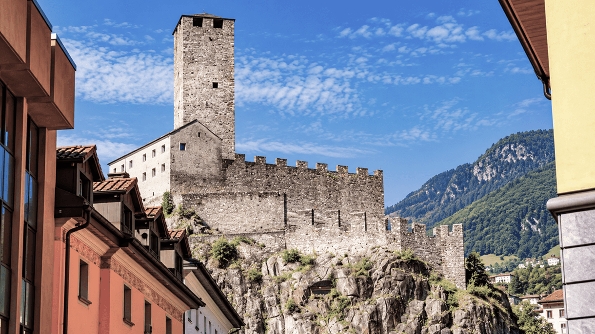 Historische Burgen von Bellinzona: Ein mittelalterlicher Steinturm mit Wehrmauern thront auf einem felsigen Hügel, eingerahmt von farbigen Stadthäusern und grünen Bergen unter blauem Himmel.