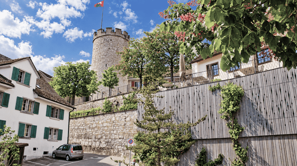 Historischer Wehrturm von Regensberg mit wehender Fahne, umgeben von blühenden Bäumen, traditionellen Häusern mit grünen Fensterläden und einer alten Natursteinmauer.
