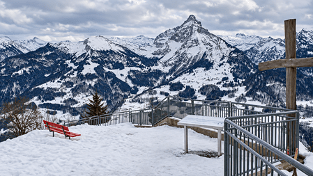 Verschneiter Aussichtspunkt mit Holzkreuz, roten Sitzbänken und Panoramablick auf schneebedeckte Schweizer Alpen unter bewölktem Himmel.