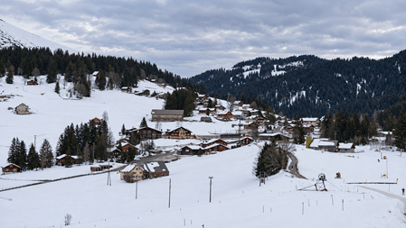 Verschneites Schweizer Bergdorf mit traditionellen Chalets, weissen Wiesen und dunklen Tannenhängen unter einem bewölkten Winterhimmel. Im Vordergrund sind ein präpariertes Schneefeld und eine kleine Skiliftanlage zu sehen.
