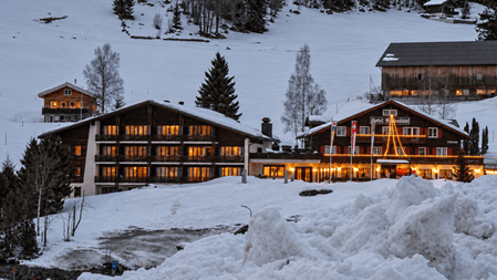 Verschneites Berghotel mit traditionellen Holzbauten, warm beleuchteten Fenstern und festlicher Lichterdekoration. Im Vordergrund liegt aufgeschobener Schnee, dahinter erstrecken sich verschneite Hänge mit Tannen und Alphütten.
