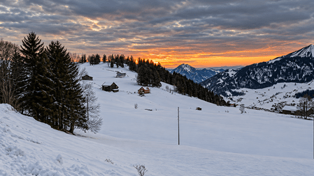 Verschneite Hügellandschaft mit verstreuten Alphütten, dunklen Tannen und schneebedeckten Bergen im Hintergrund. Orangefarbenes Abendlicht leuchtet durch die Wolken über dem winterlichen Alpental.